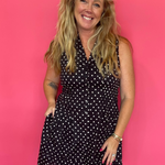A smiling blonde woman wearing a black sleeveless mini dress with white polka dots, standing against a solid bright pink studio backdrop.