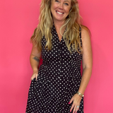 A smiling blonde woman wearing a black sleeveless mini dress with white polka dots, standing against a solid bright pink studio backdrop.