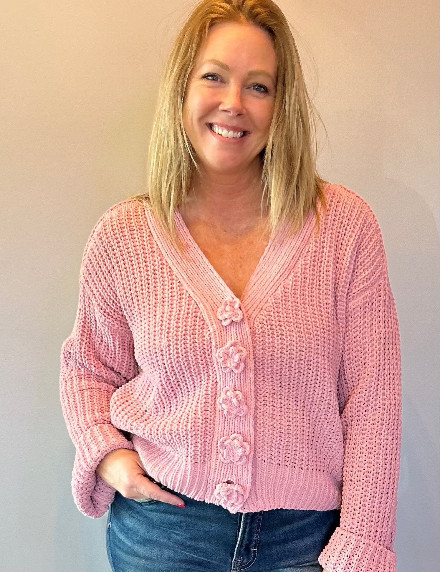 Woman wearing a pink cardigan and blue jeans standing against a plain wall.