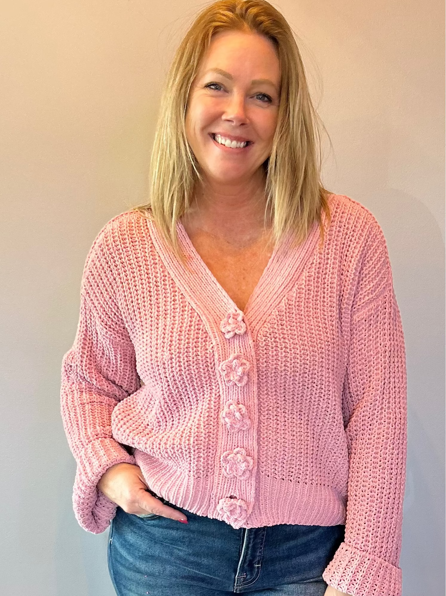 Woman wearing a pink cardigan and blue jeans standing against a plain wall.