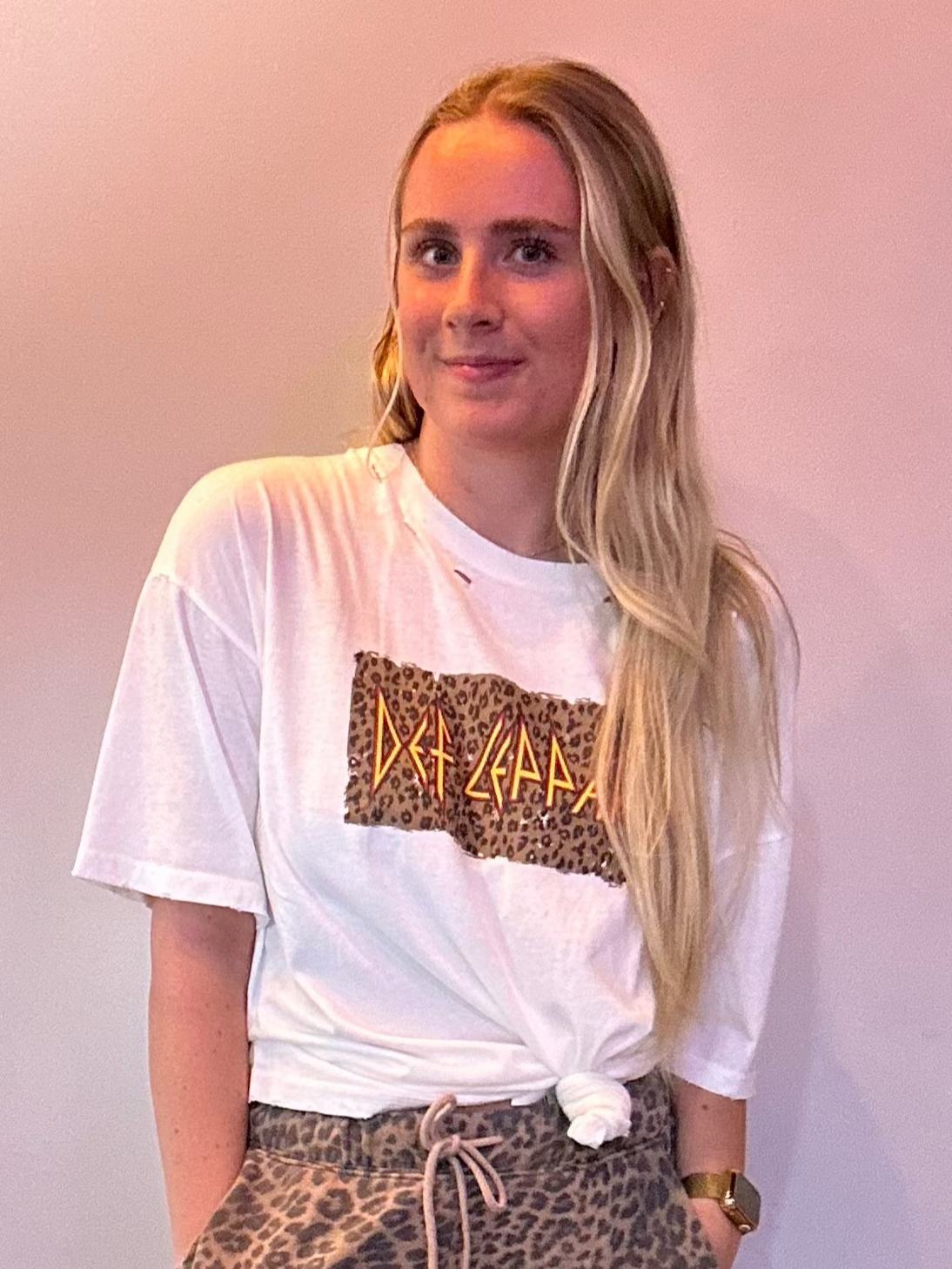 Person wearing a white t-shirt with a graphic design and leopard print pants against a plain wall.