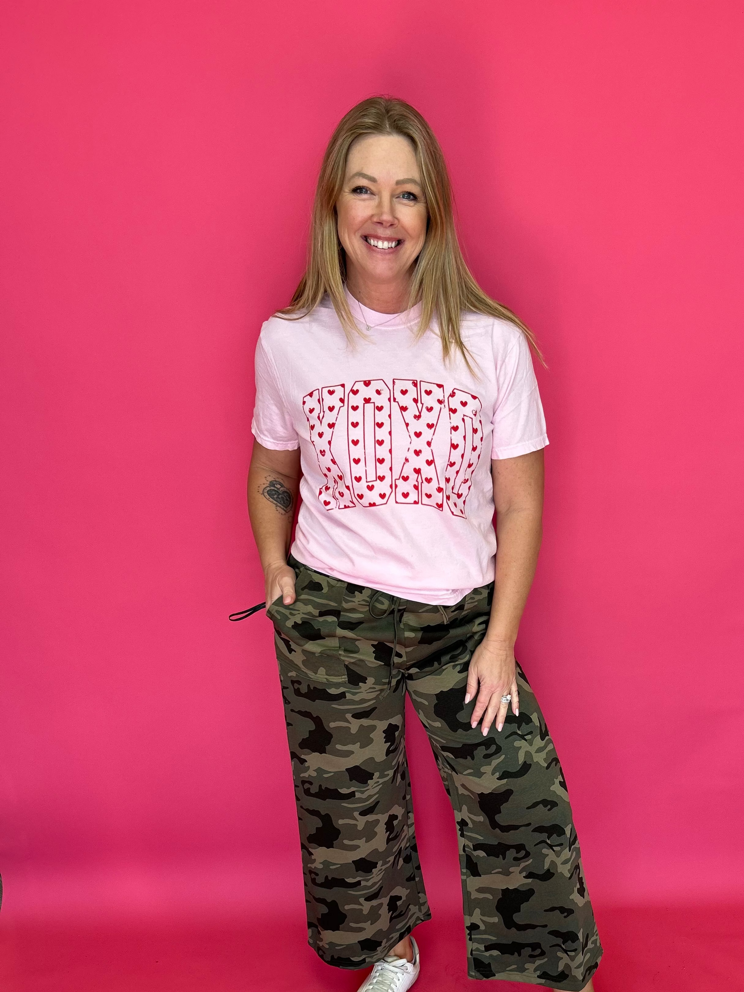 Person wearing a pink t-shirt with a red pattern and camouflage pants on a pink background