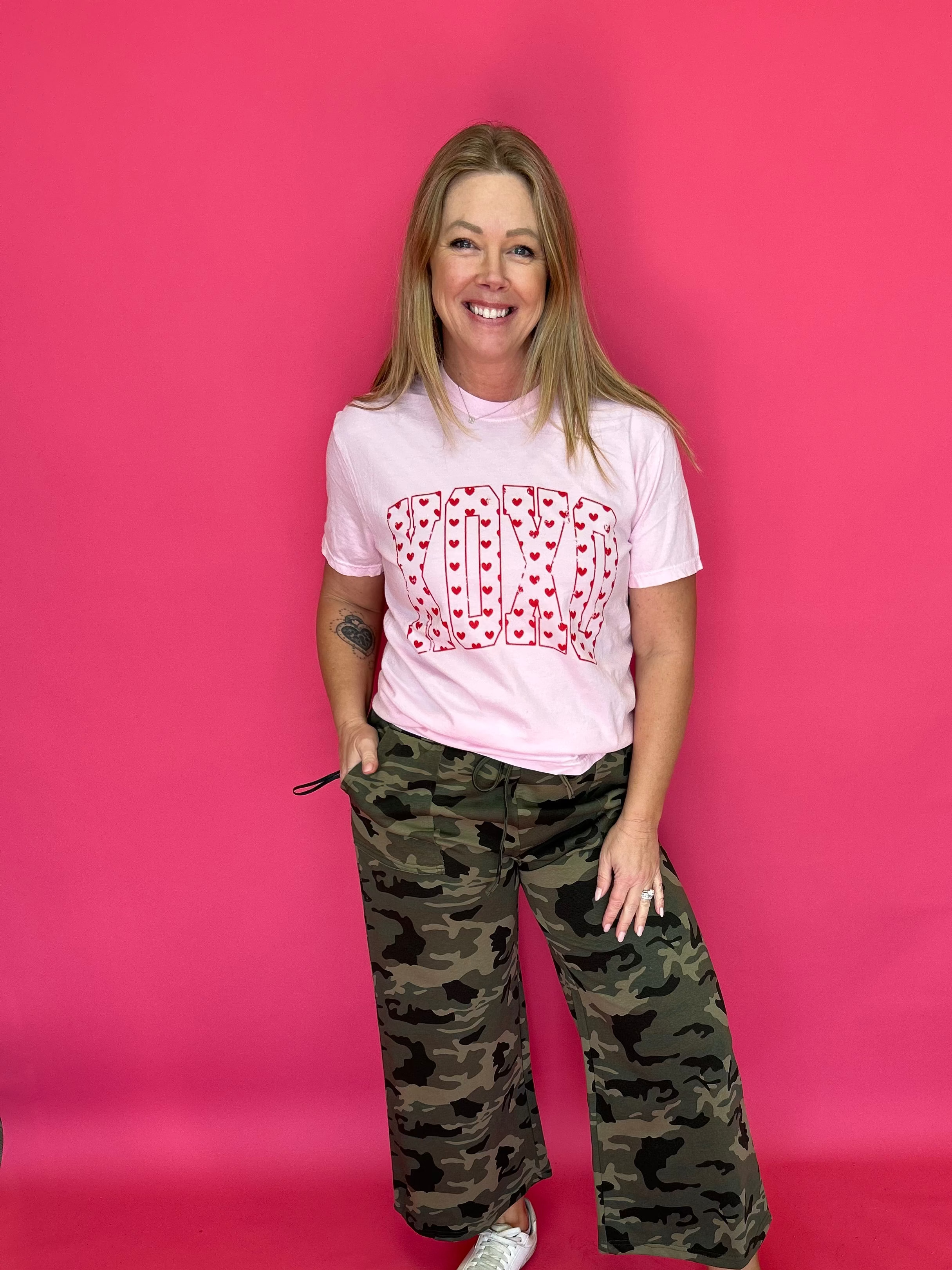 Person wearing a pink t-shirt with a red pattern and camouflage pants on a pink background