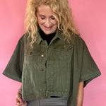 A woman with blonde curly hair wearing an olive green short-sleeve cropped denim jacket, gray pants, and a black top, standing against a solid pink studio backdrop.