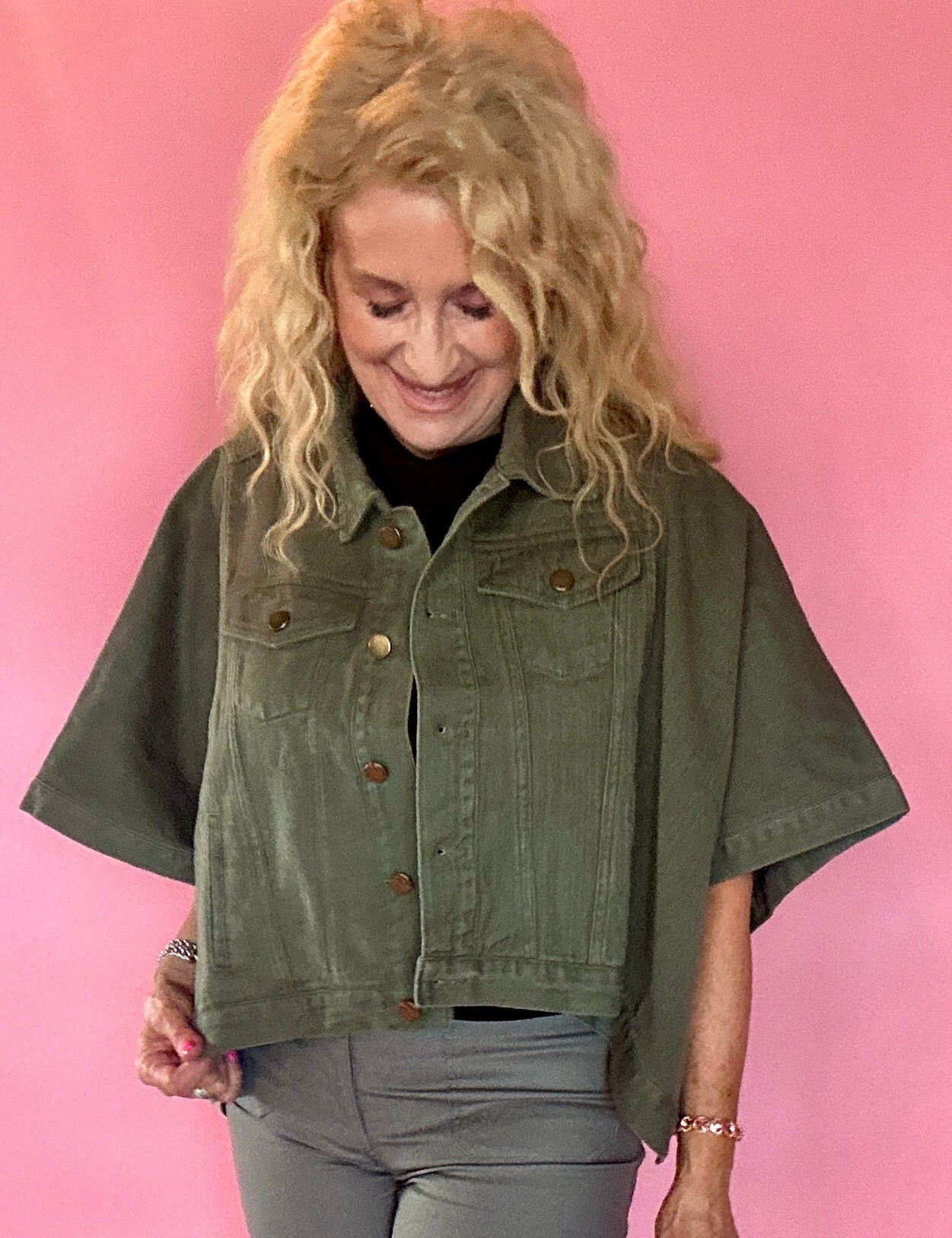 A woman with blonde curly hair wearing an olive green short-sleeve cropped denim jacket, gray pants, and a black top, standing against a solid pink studio backdrop.