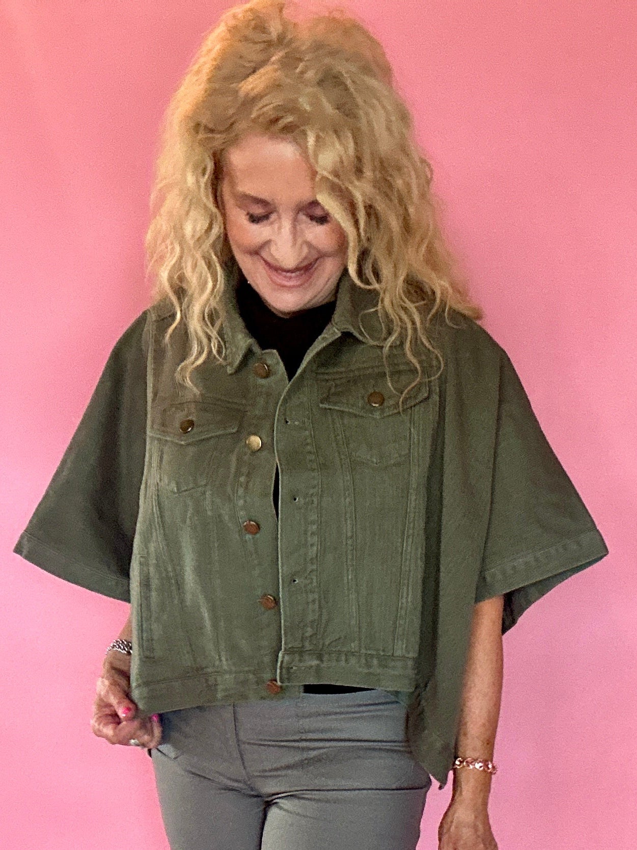 A woman with blonde curly hair wearing an olive green short-sleeve cropped denim jacket, gray pants, and a black top, standing against a solid pink studio backdrop.